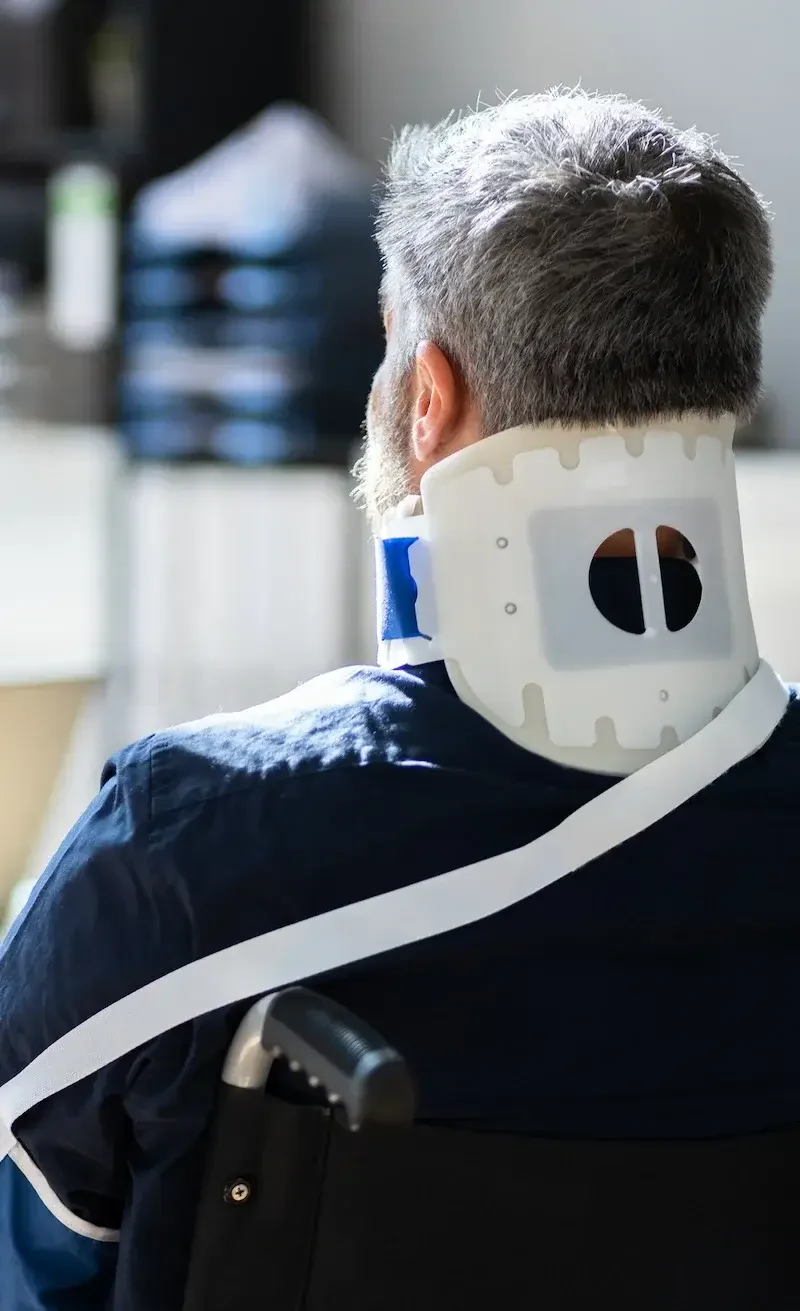 A man with gray hair wears a neck brace while seated in a wheelchair, dressed in a navy shirt, facing away from the camera.
