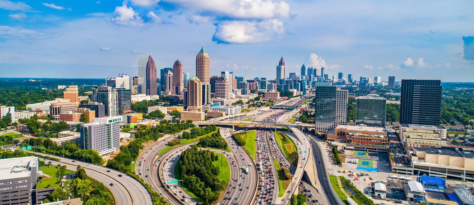 Aerial view of Atlanta's skyline with skyscrapers, highways, and greenery under a blue sky with fluffy clouds.